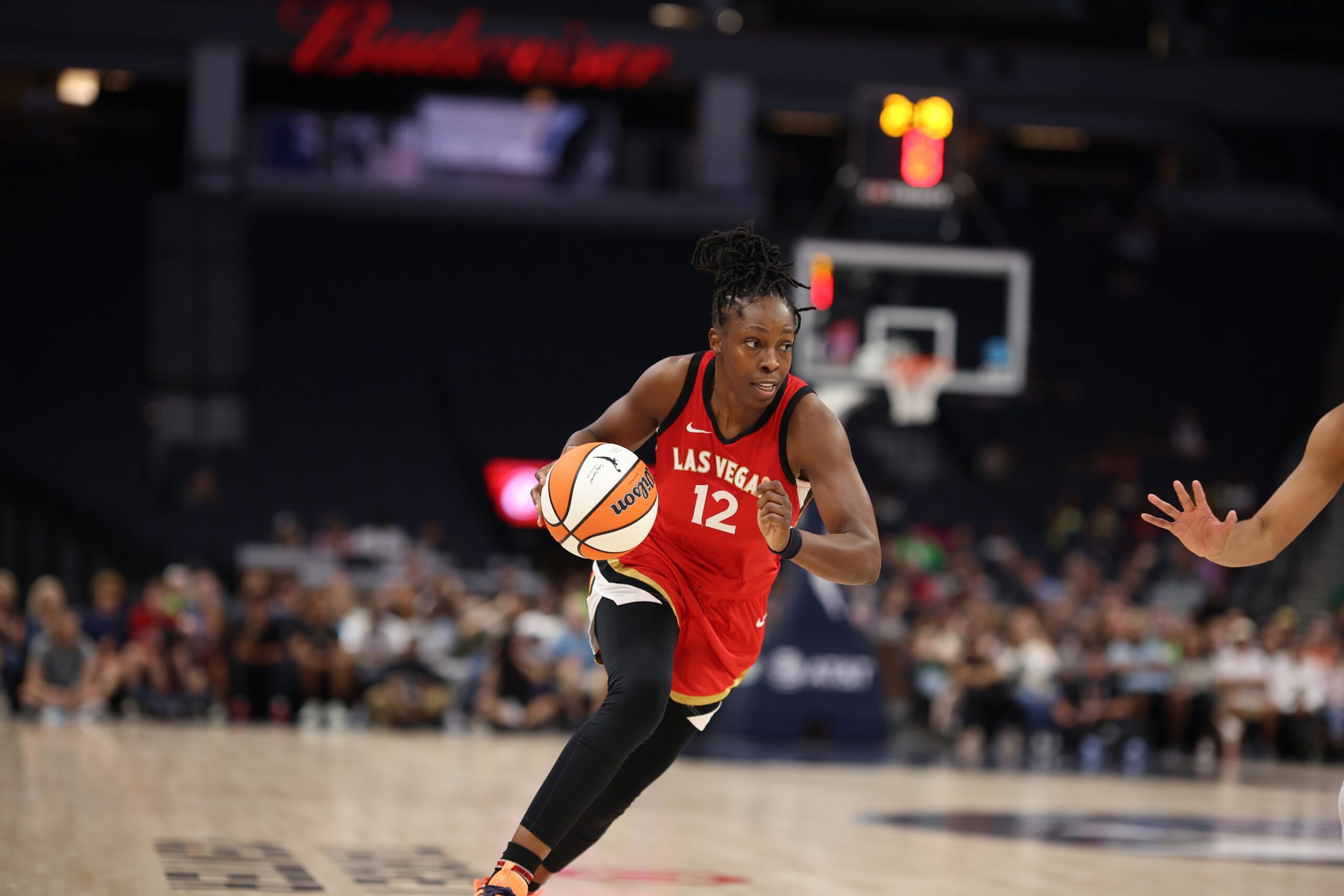 Chelsea Gray of the Las Vegas Aces drives with the ball in a road jersey during a game, with a blurred arena crowd behind her.