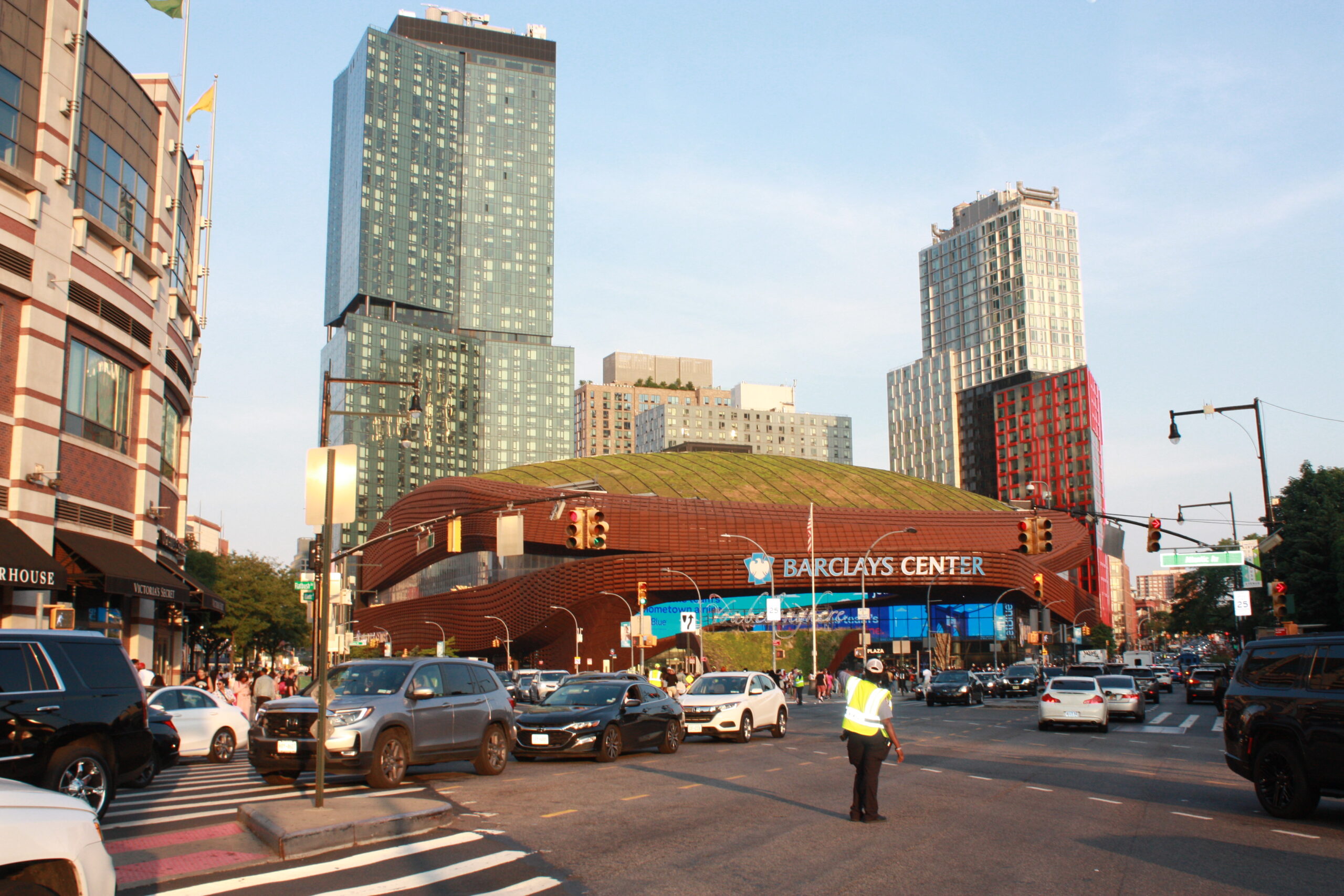 Traffic and pedestrians at the Atlantic Avenue intersection outside Barclays Center in Brooklyn, the arena's rust-coloured facade and green roof behind them