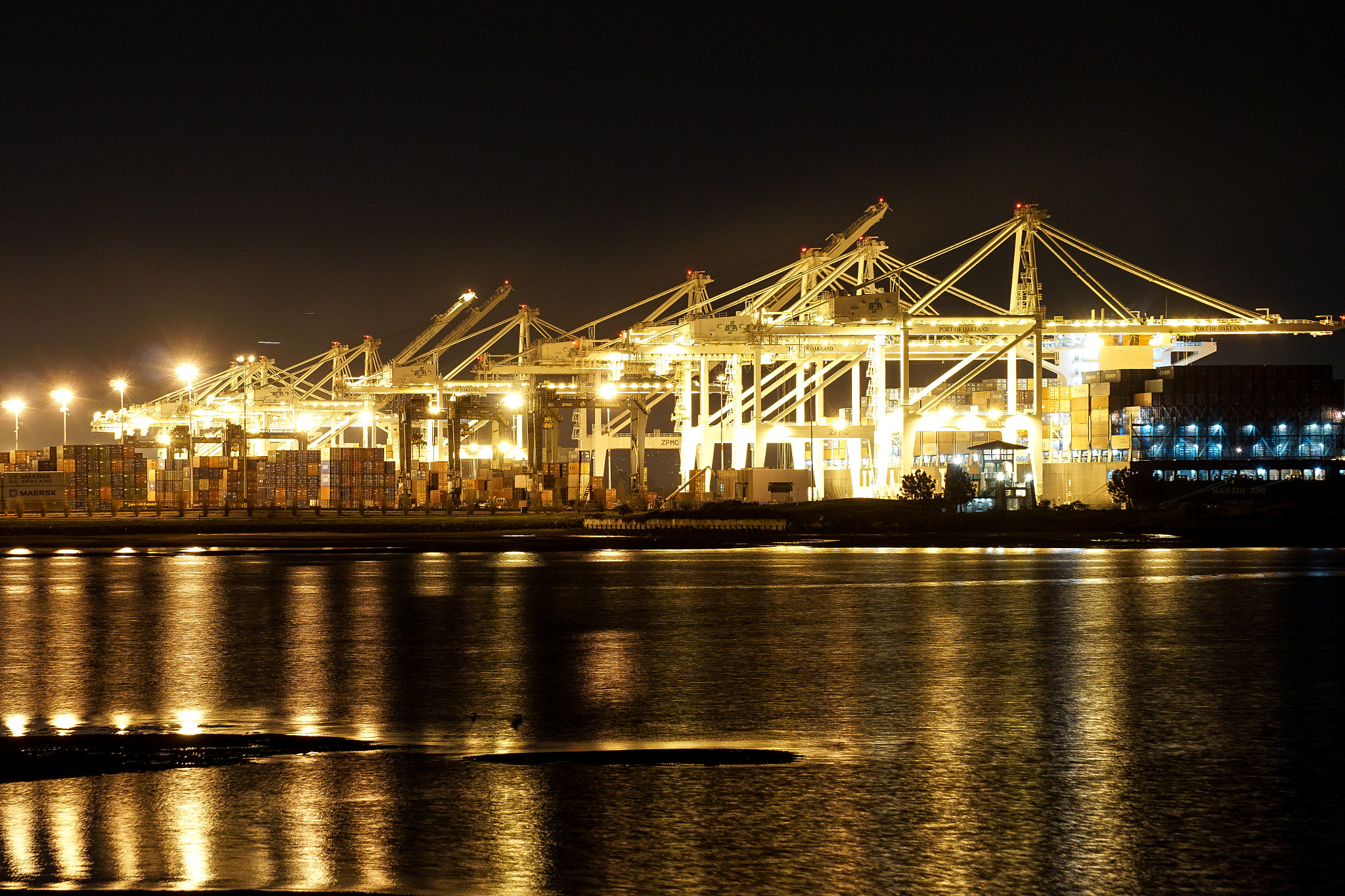 Container cranes and stacked shipping containers lit at night at the Port of Oakland, with reflections on the water in the foreground