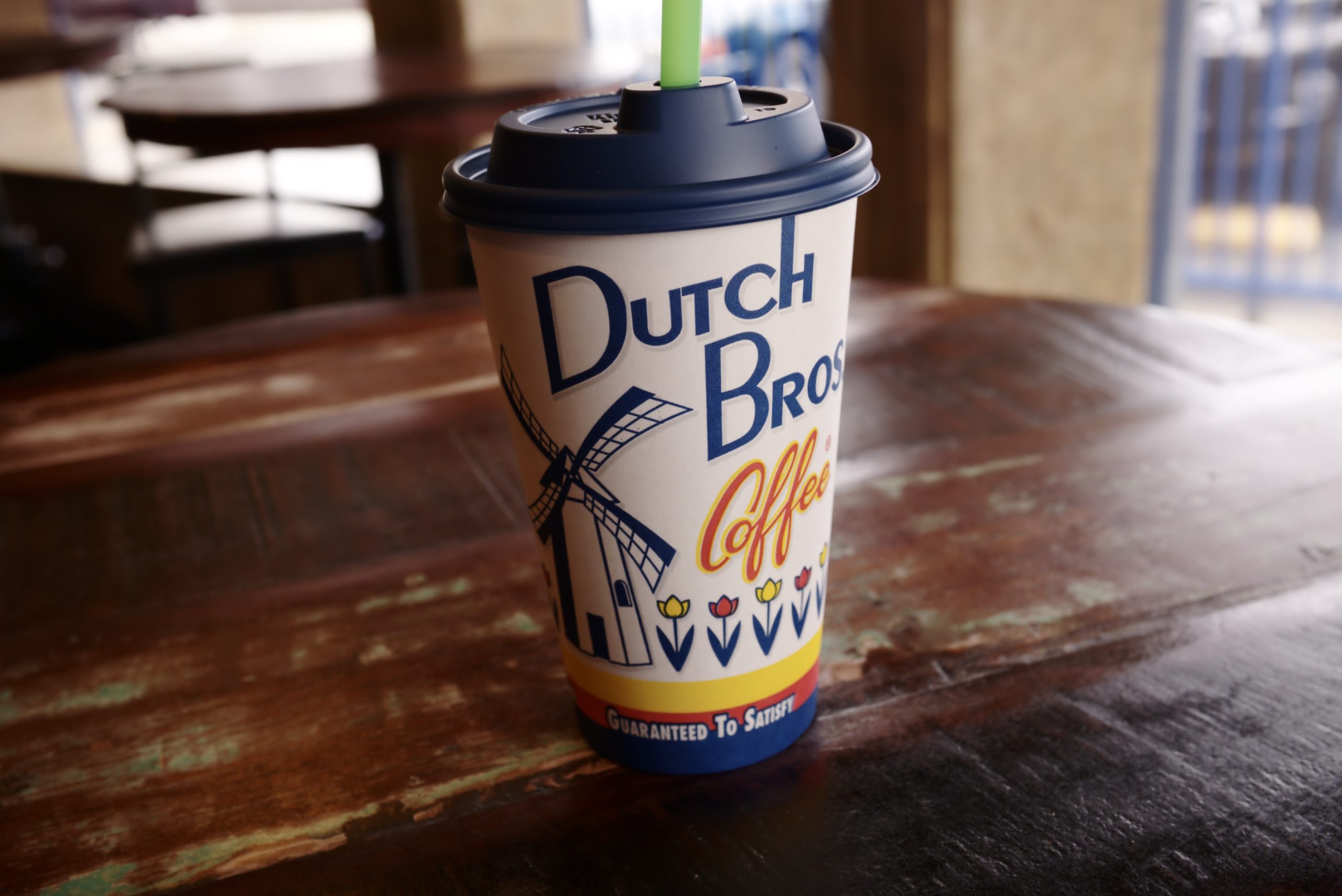 A Dutch Bros Coffee paper cup with a green straw stands on a scuffed wooden table, its blue windmill logo and tulip stripe facing the camera, a blurred cafe interior behind it