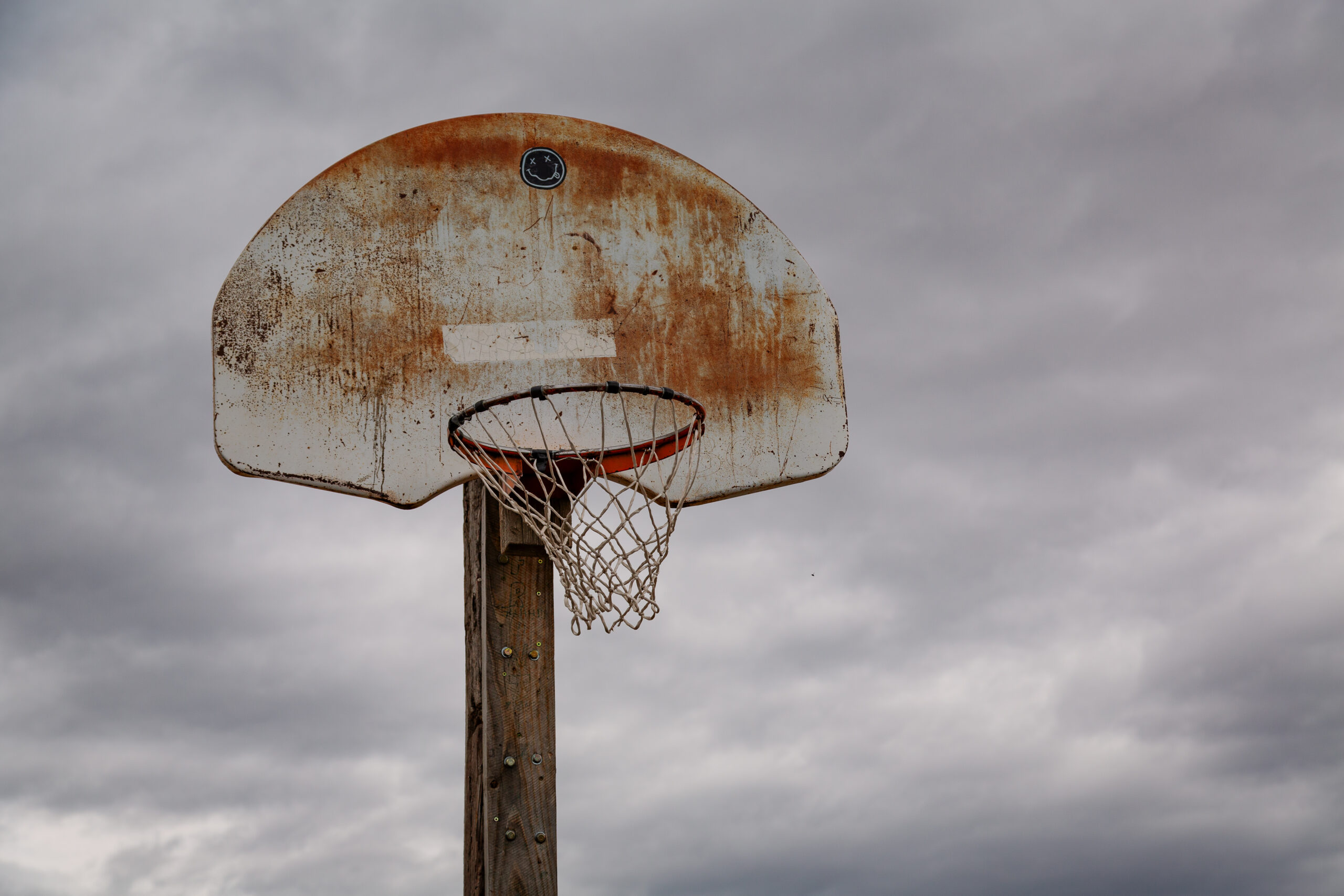 A weathered outdoor basketball backboard and rim against an overcast sky.