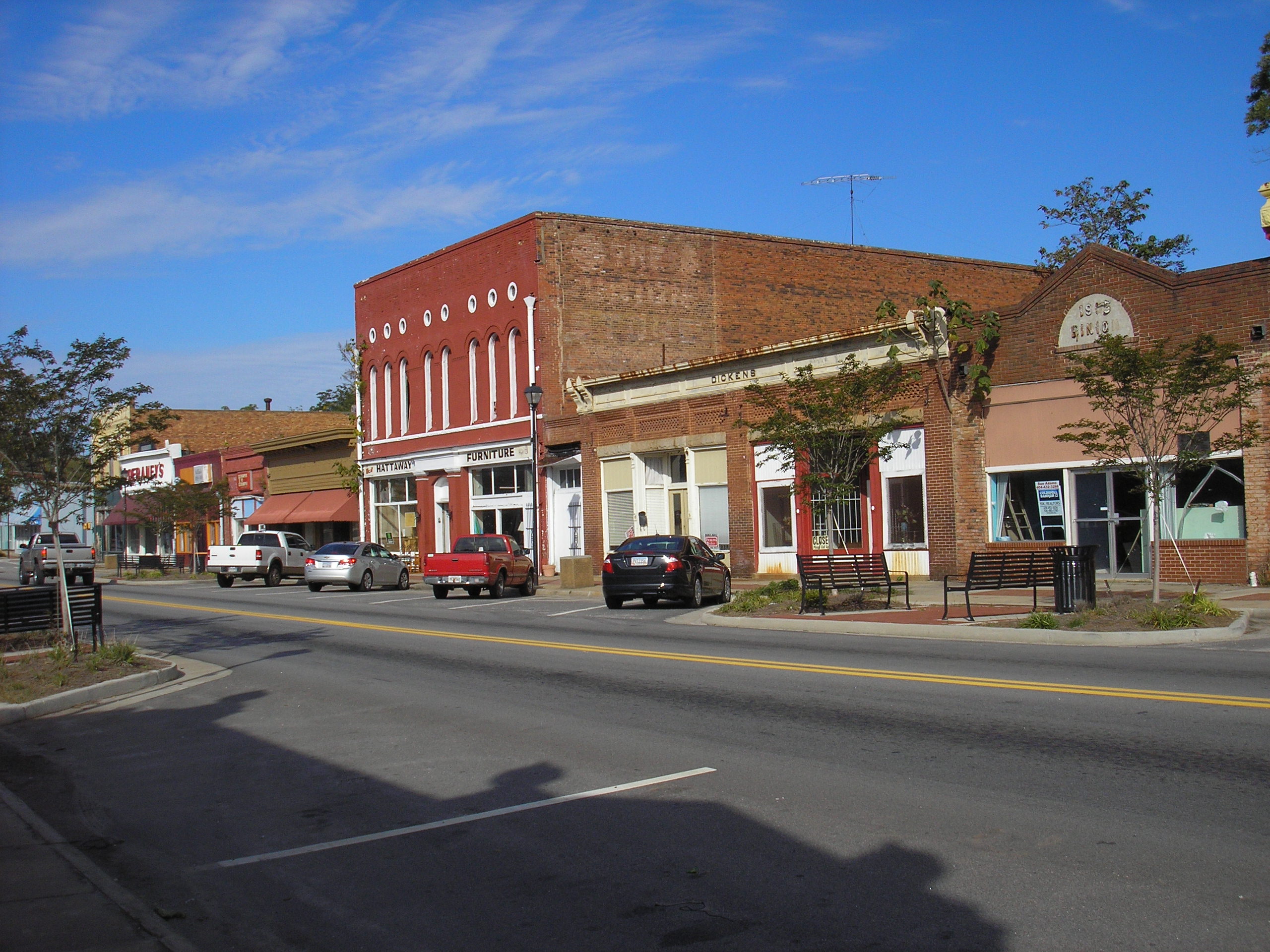 A row of brick storefronts along the main street of Sparta, Georgia, with parked cars, benches and trees in leaf under a clear sky.