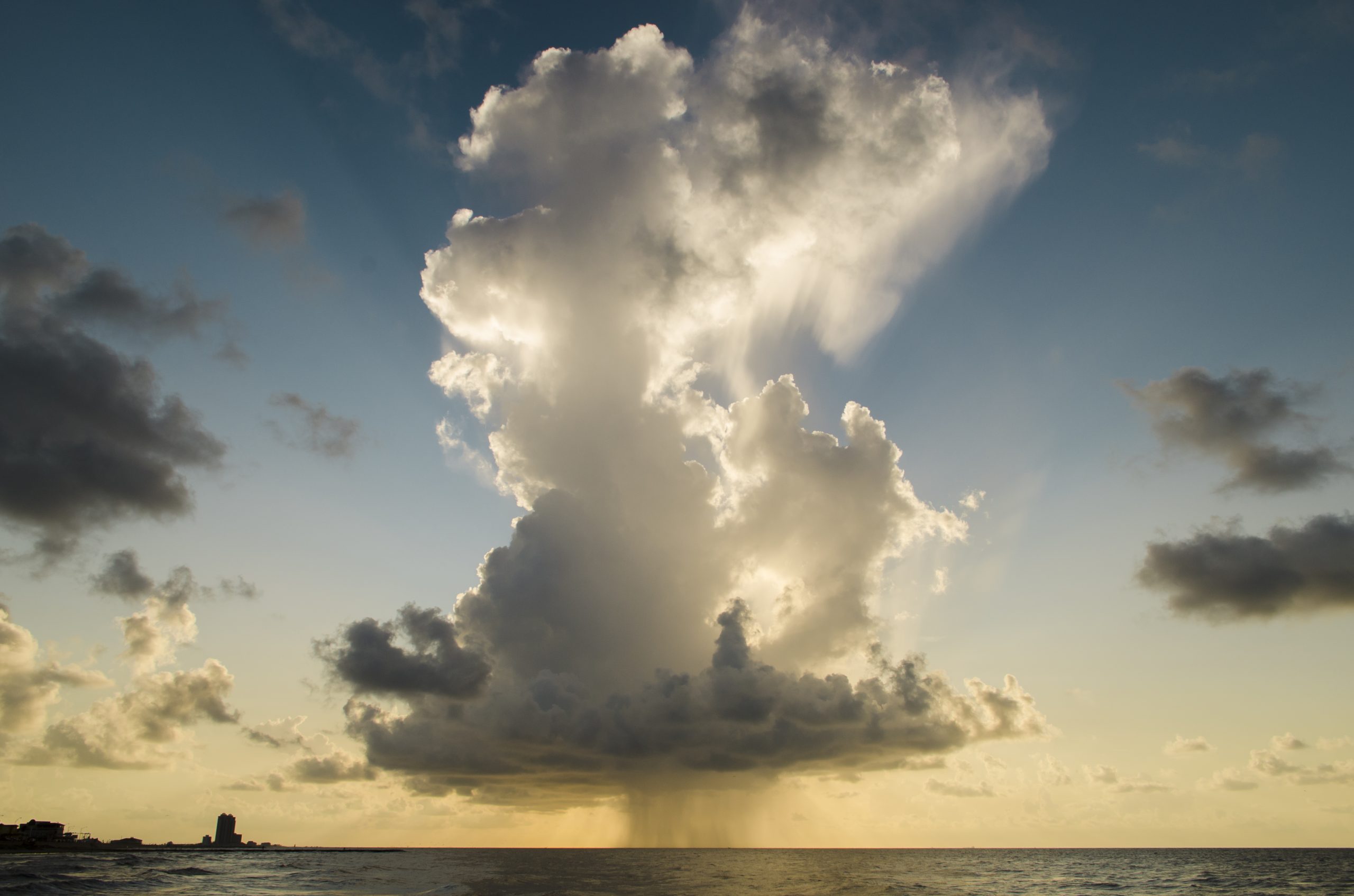 A large storm cloud with visible rainfall over the ocean near a coastline at sunset
