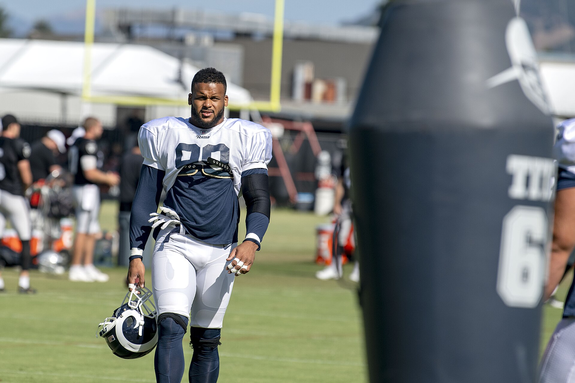 Aaron Donald in a white Rams practice jersey over a navy shirt, carrying his helmet across a practice field, with other players and a tackling dummy in the background.