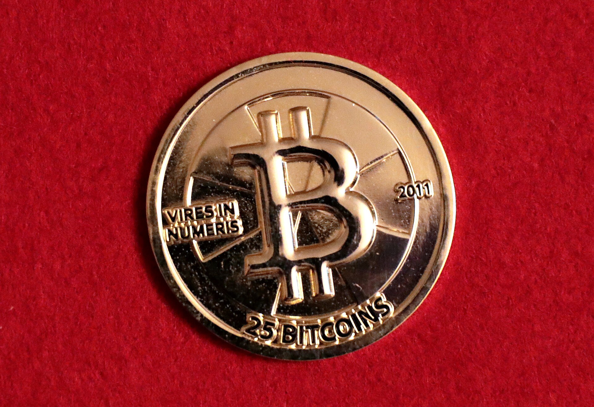 A gold-colored collectible coin stamped with the Bitcoin "B" symbol, the words "Vires in Numeris", "2011" and "25 Bitcoins", lying on red felt.