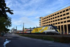 A yellow-and-gray Brightline passenger train on the tracks beside a parking garage in Fort Lauderdale, Florida.