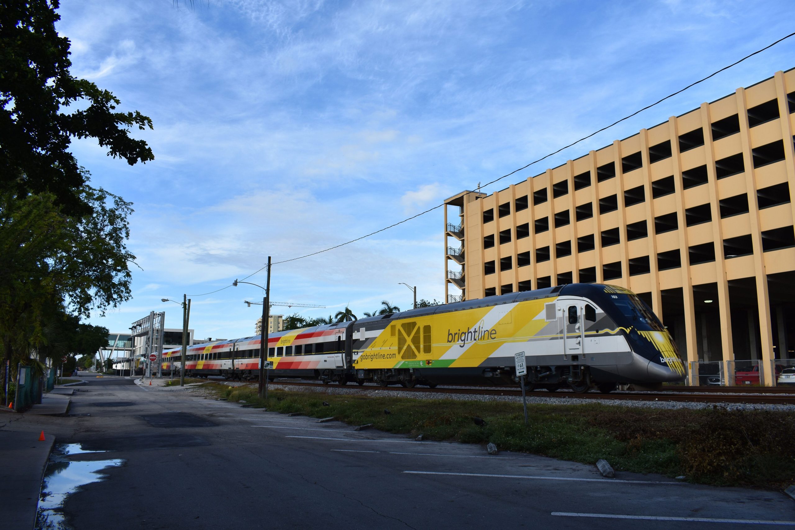 A yellow-and-gray Brightline passenger train on the tracks beside a parking garage in Fort Lauderdale, Florida.