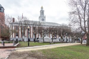 A brick and white-columned civic building with a white clock tower, seen across a park with lampposts, benches and leafless trees under an overcast sky.