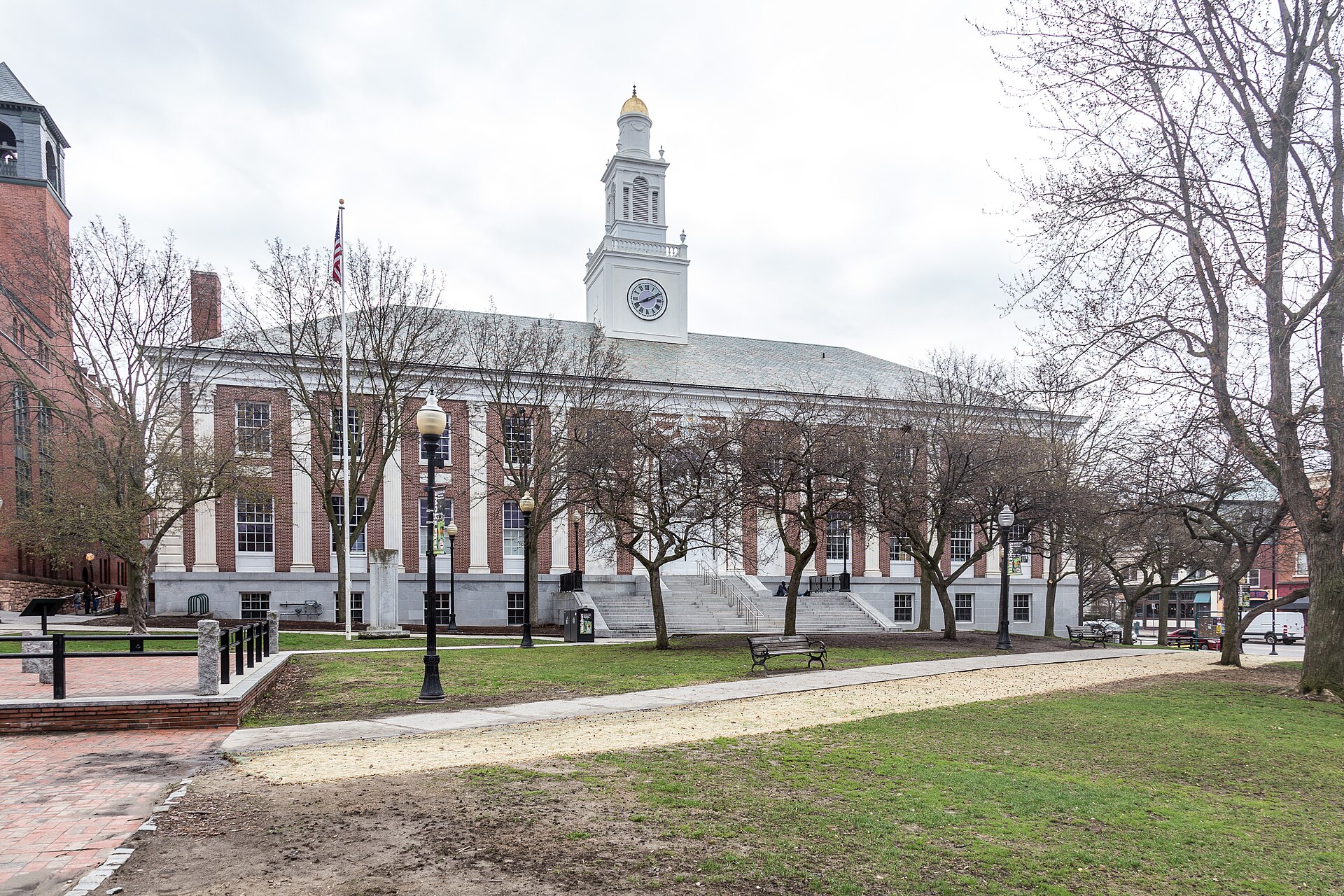 A brick and white-columned civic building with a white clock tower, seen across a park with lampposts, benches and leafless trees under an overcast sky.