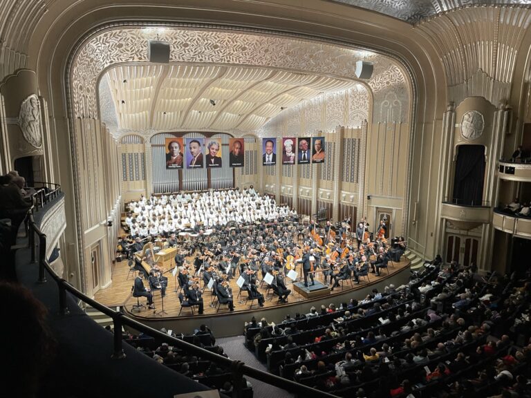 The Cleveland Orchestra and a chorus on stage in the ornate concert hall at Severance, seen from the balcony above a seated audience.