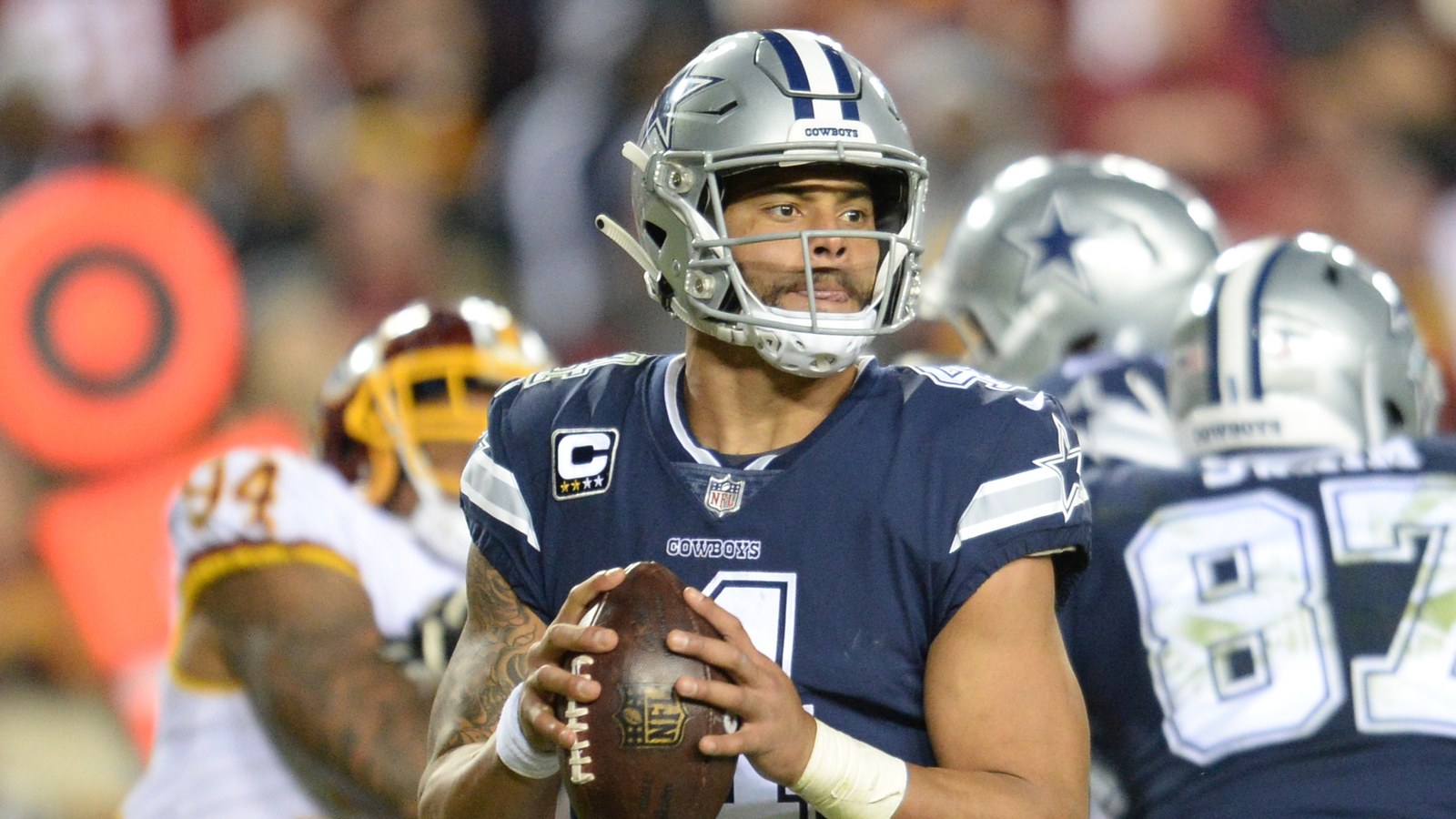 Dak Prescott in a Dallas Cowboys navy jersey and silver helmet, holding a football in both hands as he looks downfield in the pocket, with a blurred defender in white and burgundy behind him.