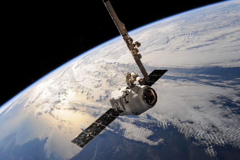 A spacecraft with deployed solar panels and a robotic arm orbits above Earth's cloud-covered surface.