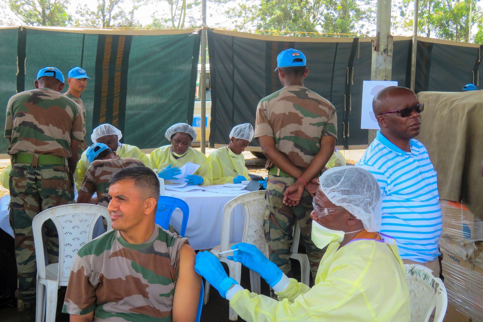 A health worker in a yellow gown, hairnet, goggles, mask and blue gloves injects an Ebola vaccine into the upper arm of a seated United Nations peacekeeper at an open-air vaccination point in the Democratic Republic of the Congo, with more health workers filling out paperwork at a table behind them.