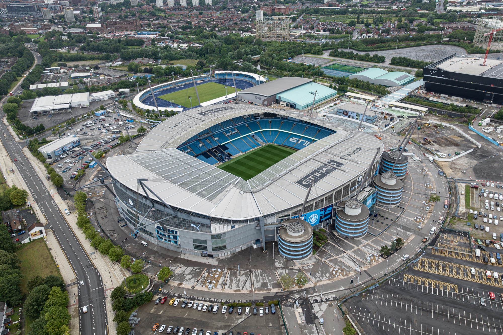 Aerial view of Manchester City's Etihad Stadium in Manchester, England, with its white roof, sky-blue seats and green pitch, surrounded by car parks and a second smaller stadium.