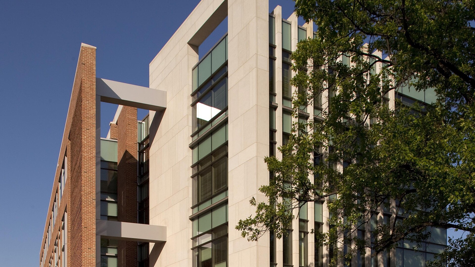 The stone-and-glass exterior of a modern government office building with a brick wing beside it, seen from a lawn under a clear blue sky, with tree branches at the right.