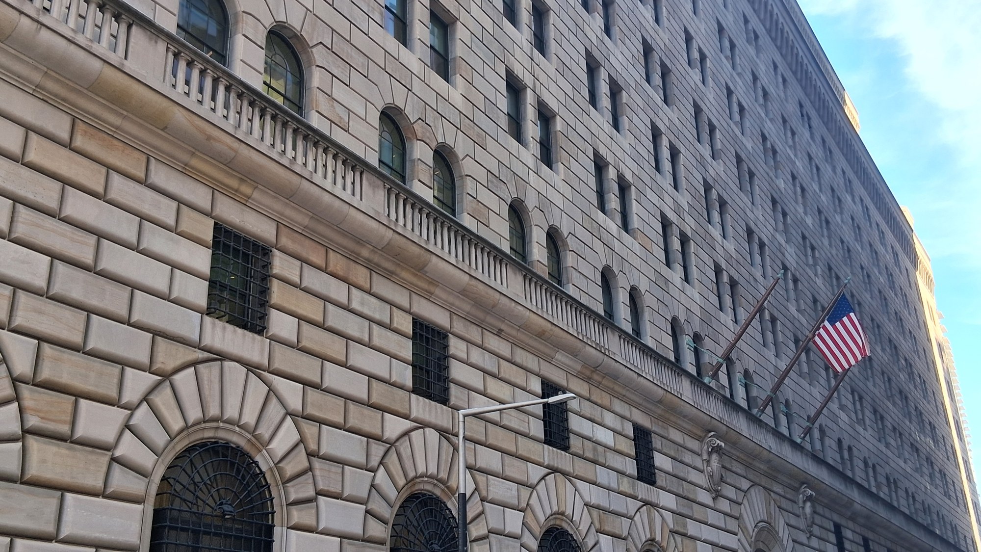 The stone facade of the Federal Reserve Bank of New York building in Lower Manhattan, with arched windows, iron grilles and an American flag.