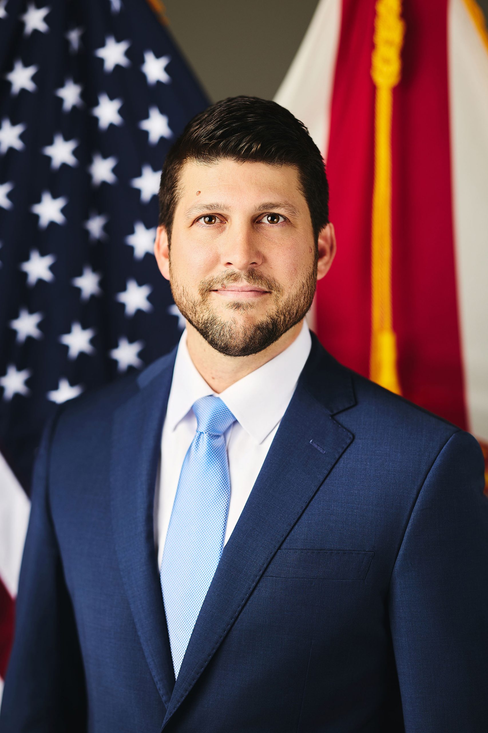 Official portrait of Florida Attorney General James Uthmeier in a navy suit and light-blue tie in front of the U.S. and Florida flags.