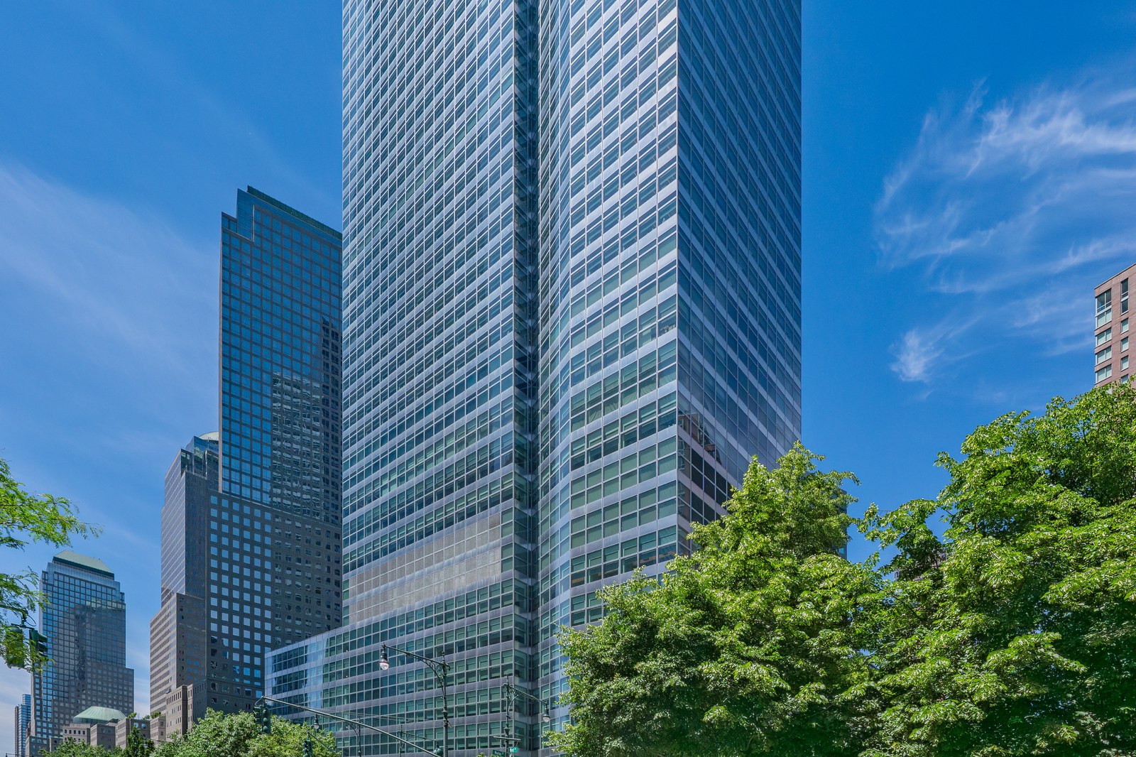 The glass tower of Goldman Sachs' headquarters at 200 West Street in Lower Manhattan under a blue sky, with leafy trees in the foreground and neighboring office towers to the left.