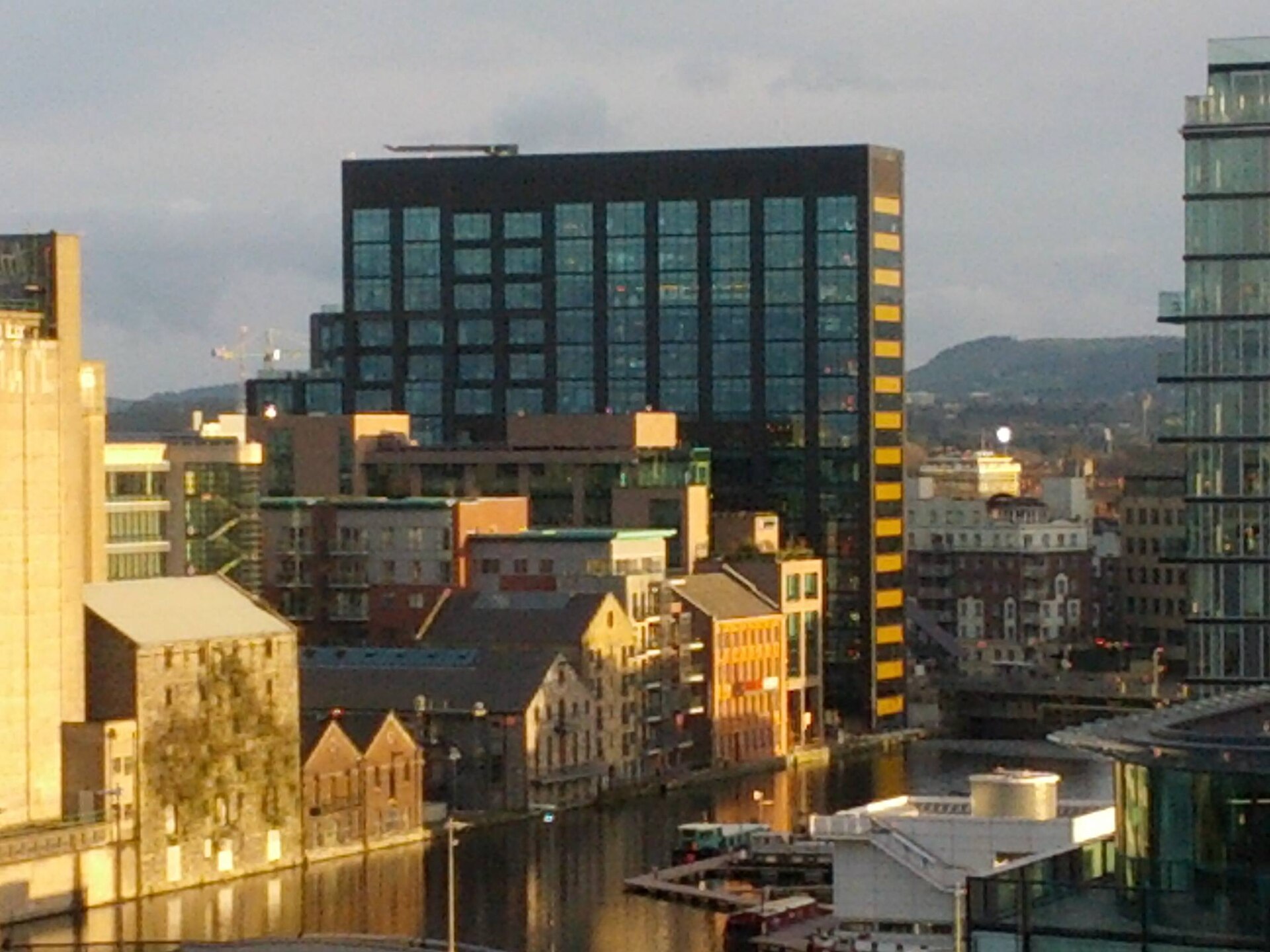 A tall dark-glass office tower with a column of yellow-banded windows on its right side, rising above older brick warehouses and apartment blocks beside a canal basin in Dublin, with hills behind.
