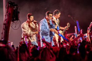 Nick, Joe and Kevin Jonas performing on stage together with microphones and a guitar.