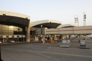 A terminal concourse at King Khalid International Airport in Riyadh, its curved roof sections running away from the camera with jet bridges attached, seen across an empty apron at dusk.