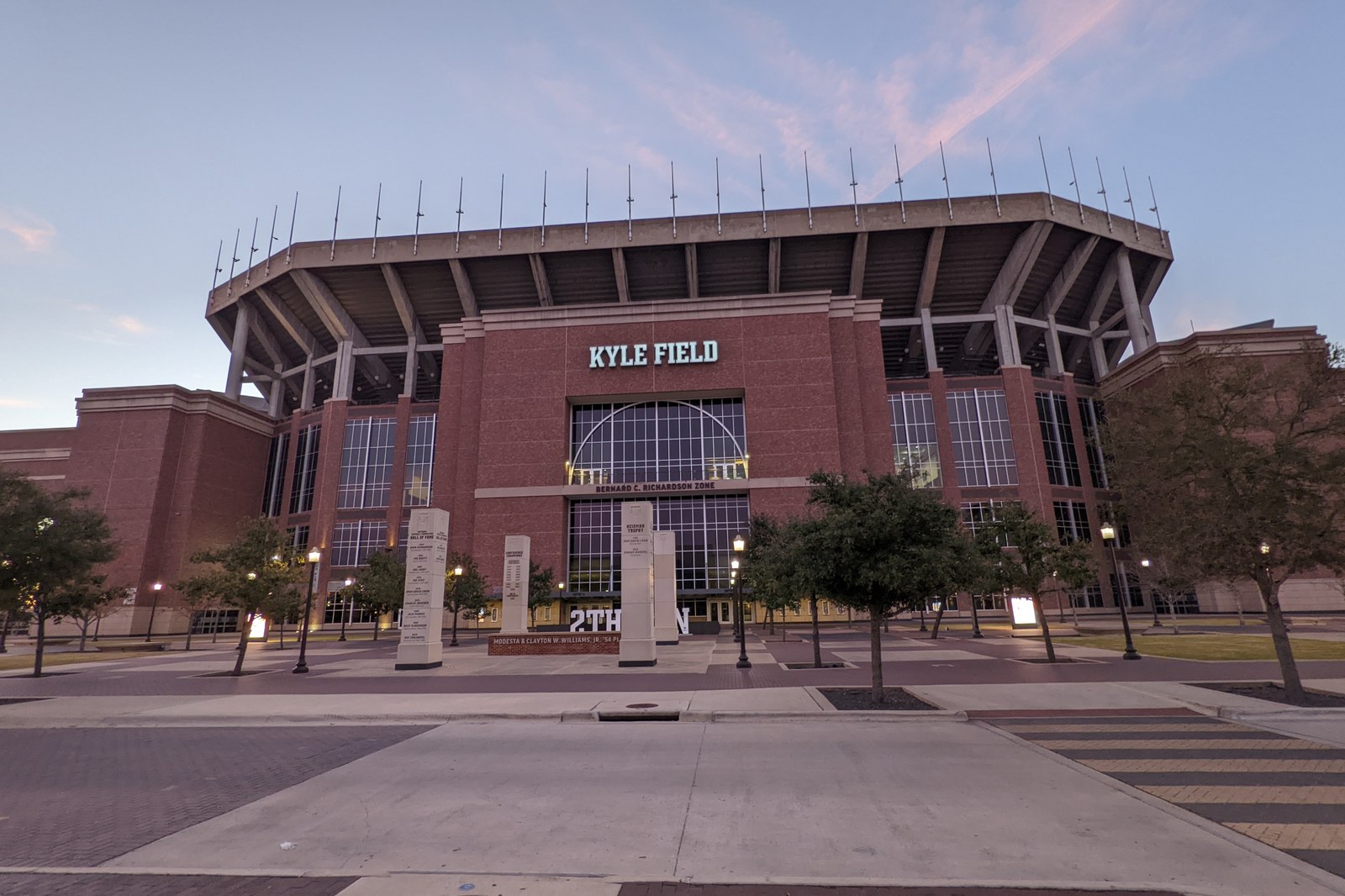 The brick exterior of Kyle Field at Texas A&M University at dusk, with the stadium name above the entrance and trees along the plaza in front.