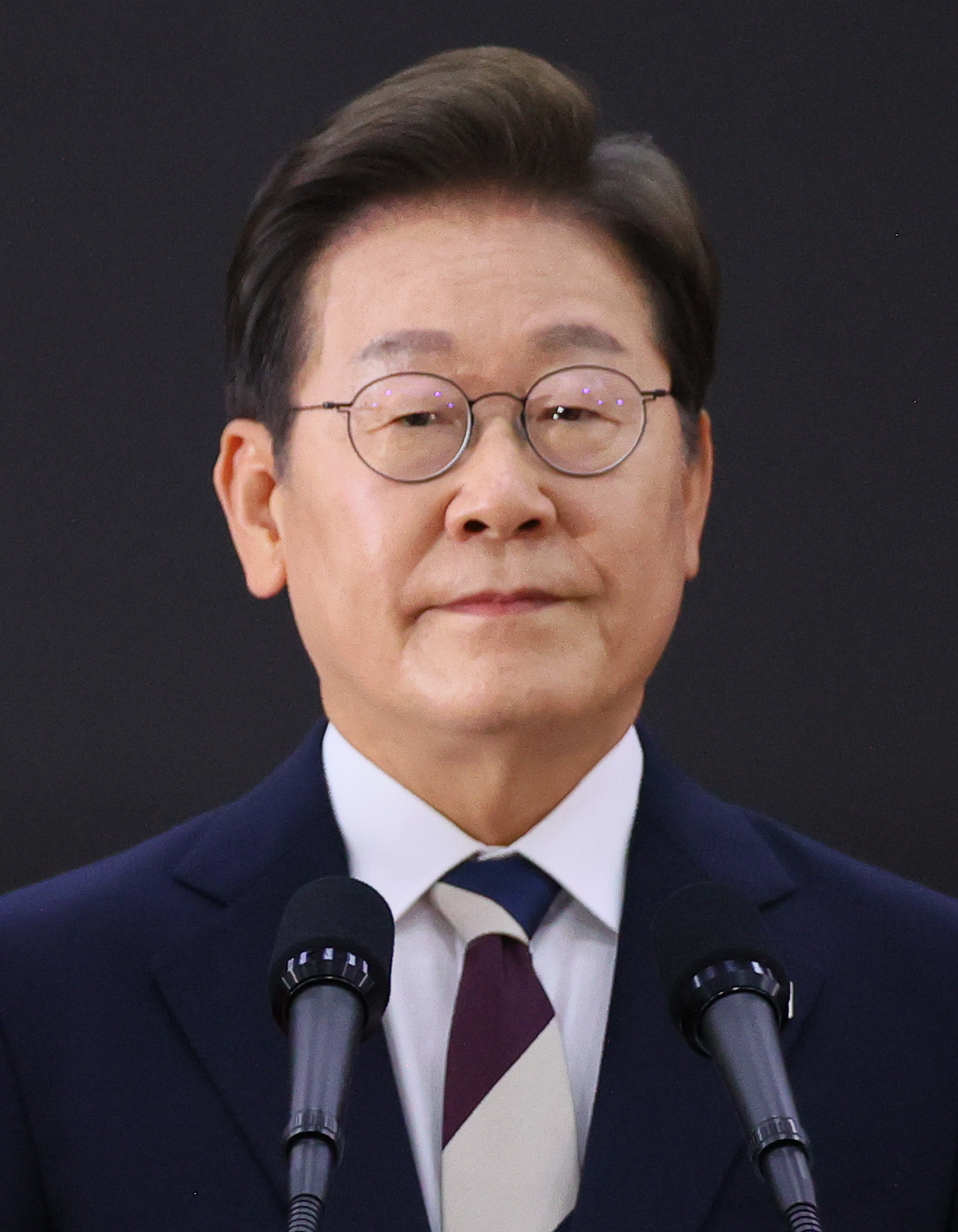 South Korean President Lee Jae-myung standing behind two microphones against a dark background.