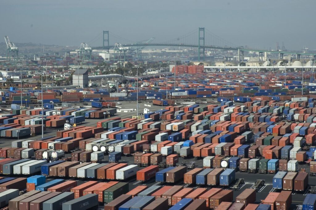 Rows of stacked shipping containers at the Port of Long Beach, California, with container cranes and a suspension bridge in the hazy background.