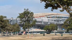 The curved, elevated form of the Lucas Museum of Narrative Art rises above scaffolding and a green construction fence in Exposition Park, Los Angeles, seen across a dry lawn and a line of trees.