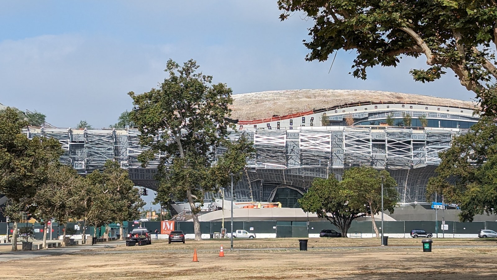 The curved, elevated form of the Lucas Museum of Narrative Art rises above scaffolding and a green construction fence in Exposition Park, Los Angeles, seen across a dry lawn and a line of trees.