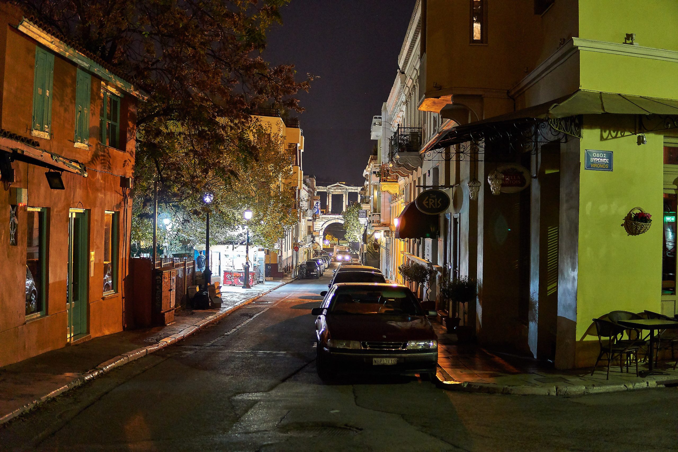Lysikratous Street in Athens' Plaka district at night, lined with low buildings and parked cars, with the Arch of Hadrian in the distance.