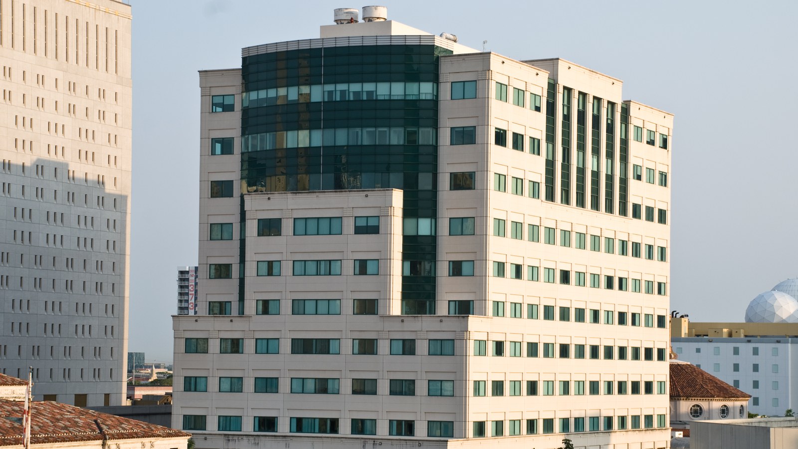 A pale stone federal courthouse tower in Miami with a curved dark-green glass section near the top, seen in late-afternoon light beside an older building.