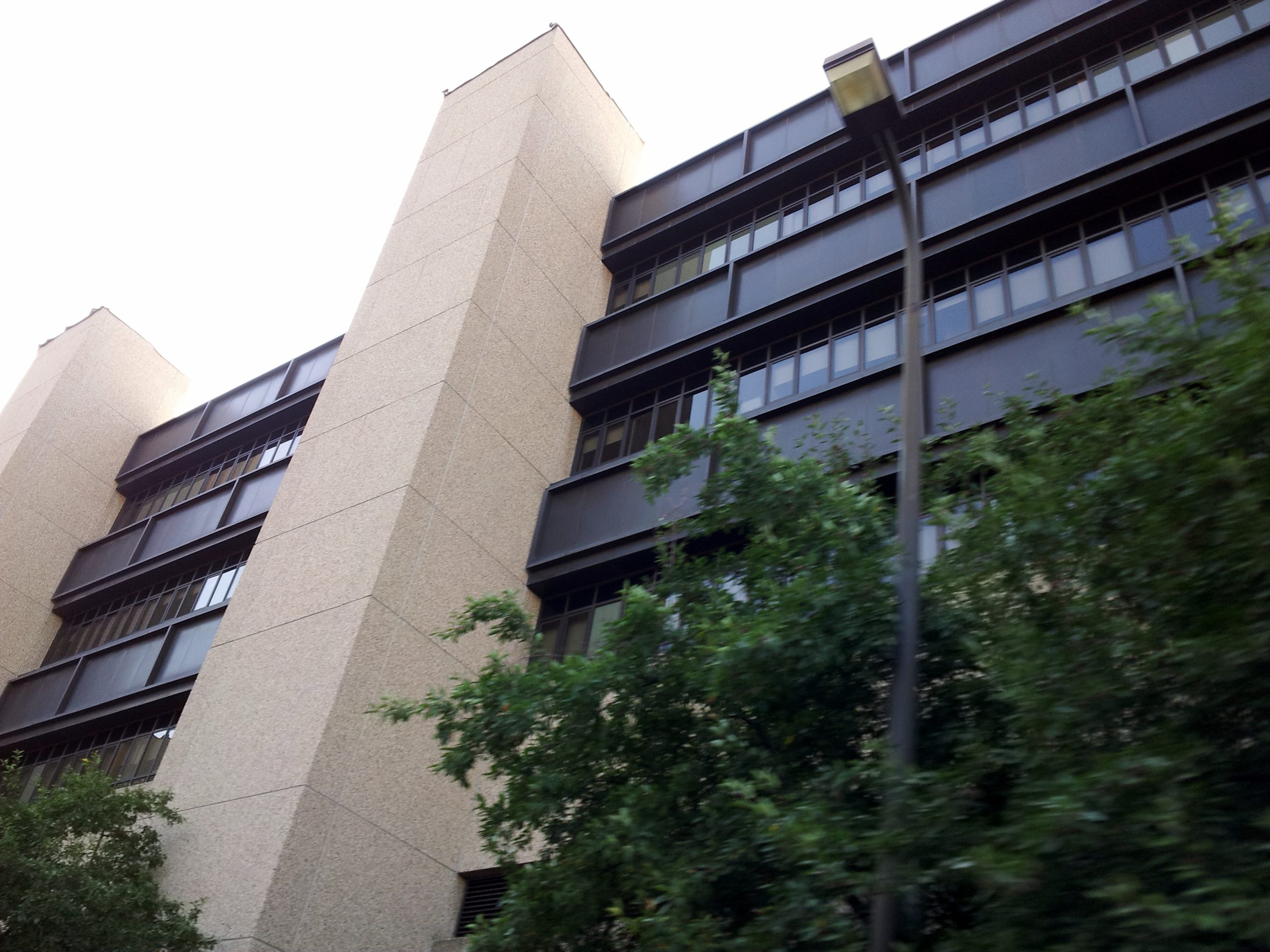 A low-angle view of the Hennepin County Medical Center building in Minneapolis, its dark window bands and pale stone columns seen from the street behind trees