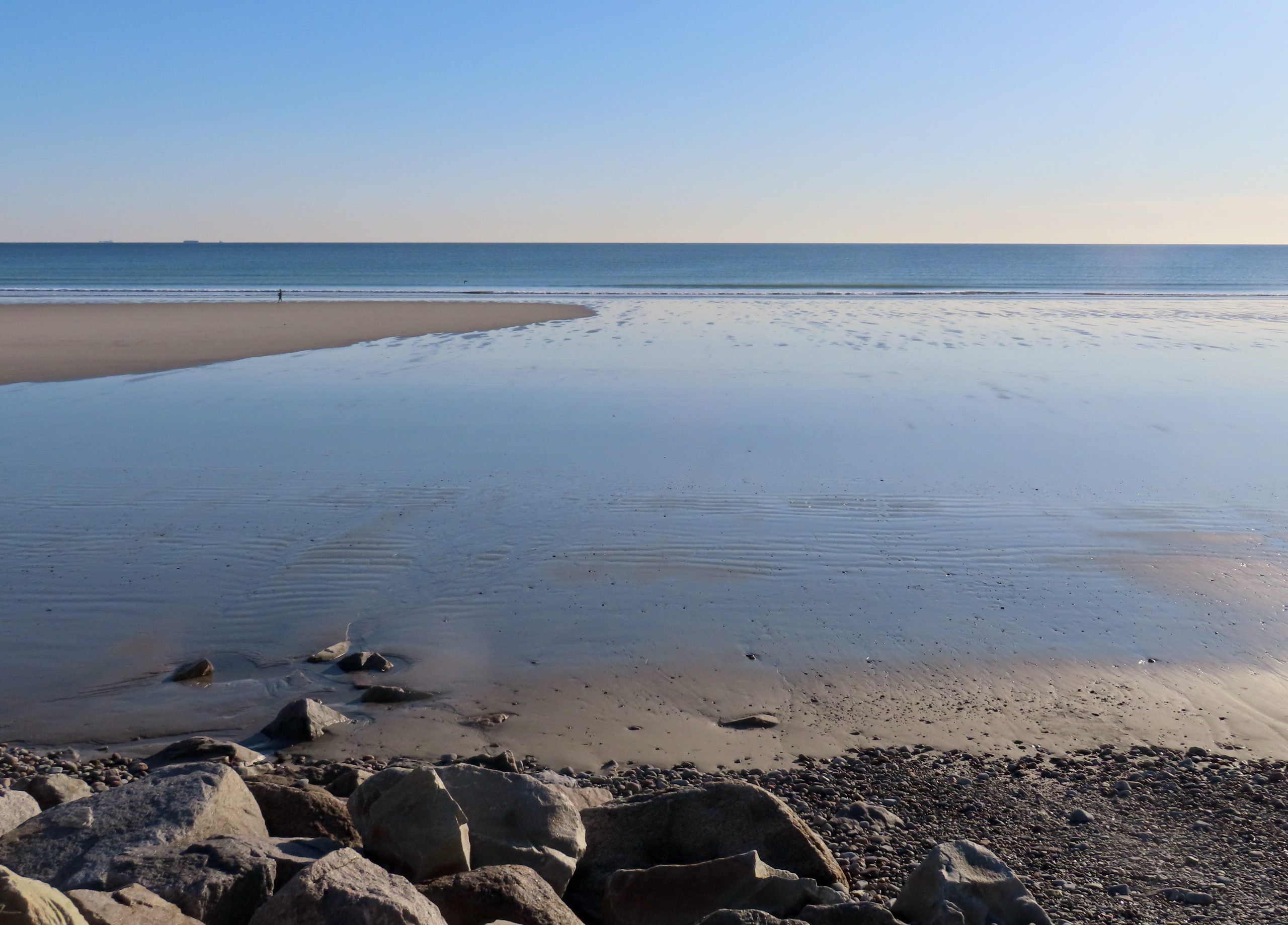 A calm, empty stretch of Nantasket Beach in Hull, Massachusetts, at low tide, with rocks in the foreground.