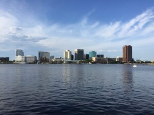 The downtown Norfolk, Virginia, skyline seen across the calm Elizabeth River under a partly cloudy blue sky.
