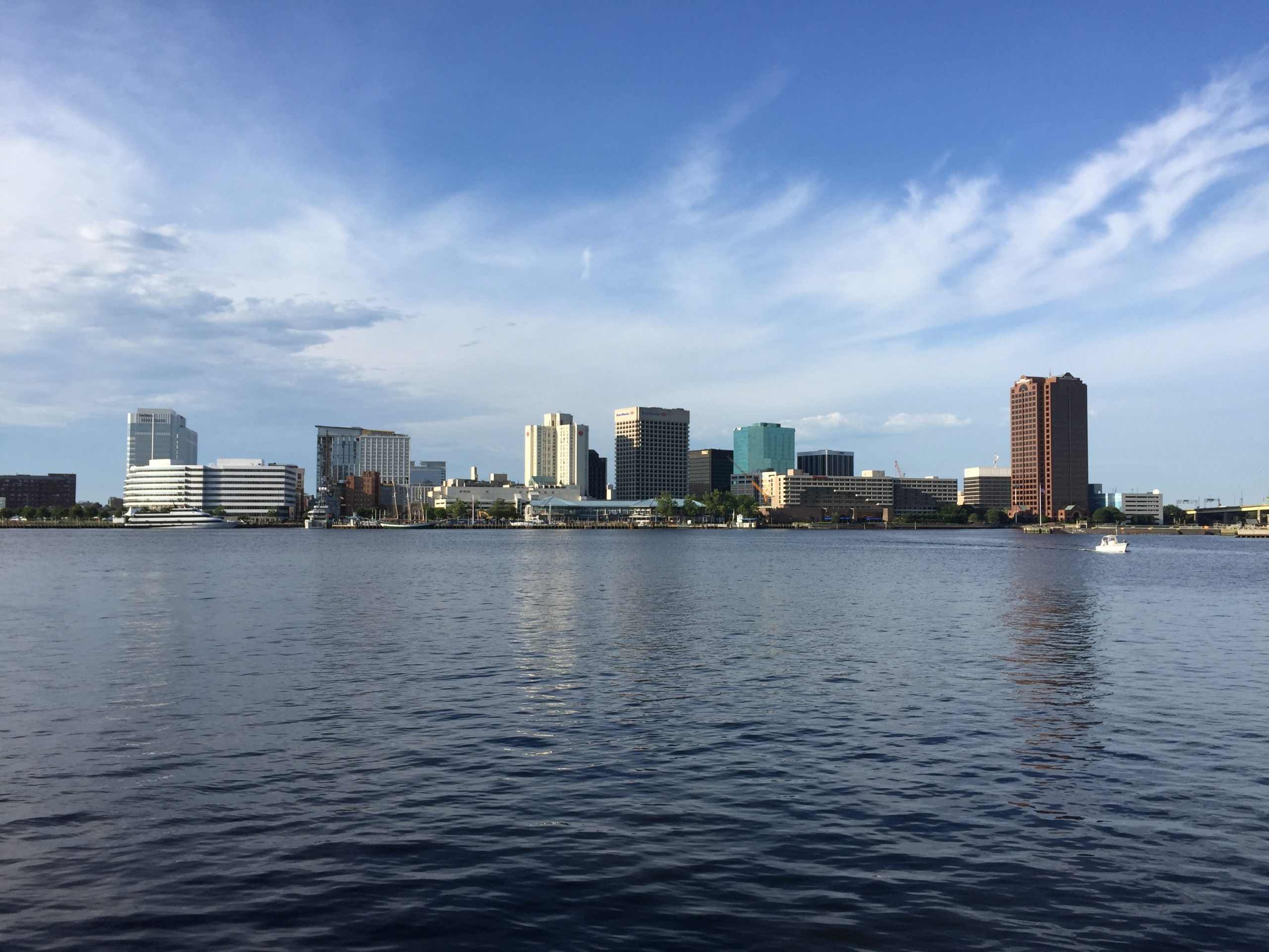 The downtown Norfolk, Virginia, skyline seen across the calm Elizabeth River under a partly cloudy blue sky.