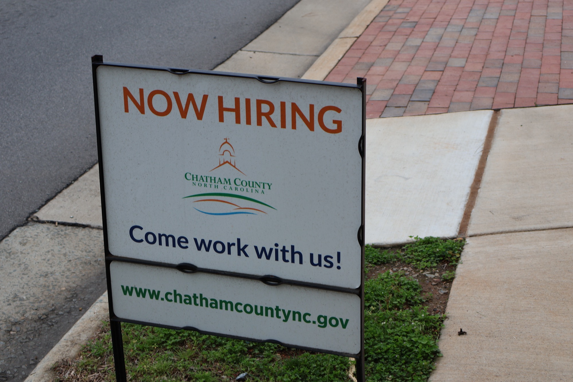 A roadside sign reading "Now Hiring — Come work with us!" for Chatham County, North Carolina, beside a brick sidewalk.
