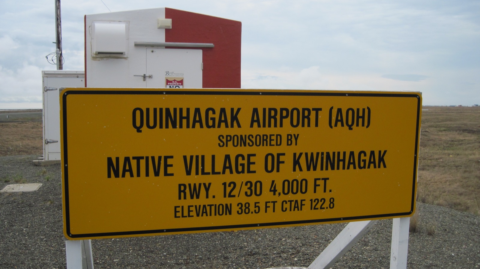 A yellow sign reading 'Quinhagak Airport (AQH), sponsored by Native Village of Kwinhagak' stands on gravel in front of flat brown tundra under a cloudy sky.