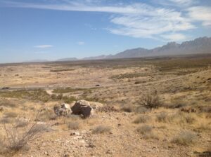 Open desert scrubland with scattered rocks and a two-lane road, leading to the jagged Organ Mountains under a blue sky near Las Cruces, New Mexico.