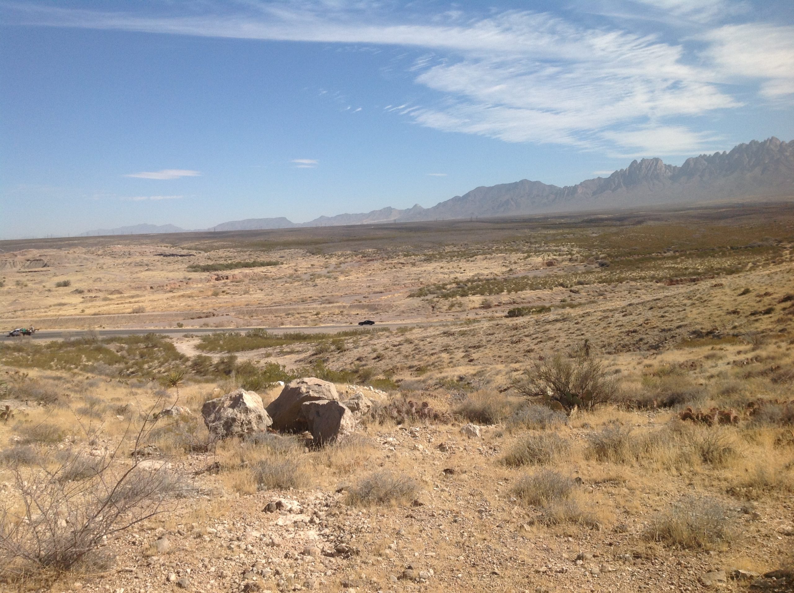 Open desert scrubland with scattered rocks and a two-lane road, leading to the jagged Organ Mountains under a blue sky near Las Cruces, New Mexico.