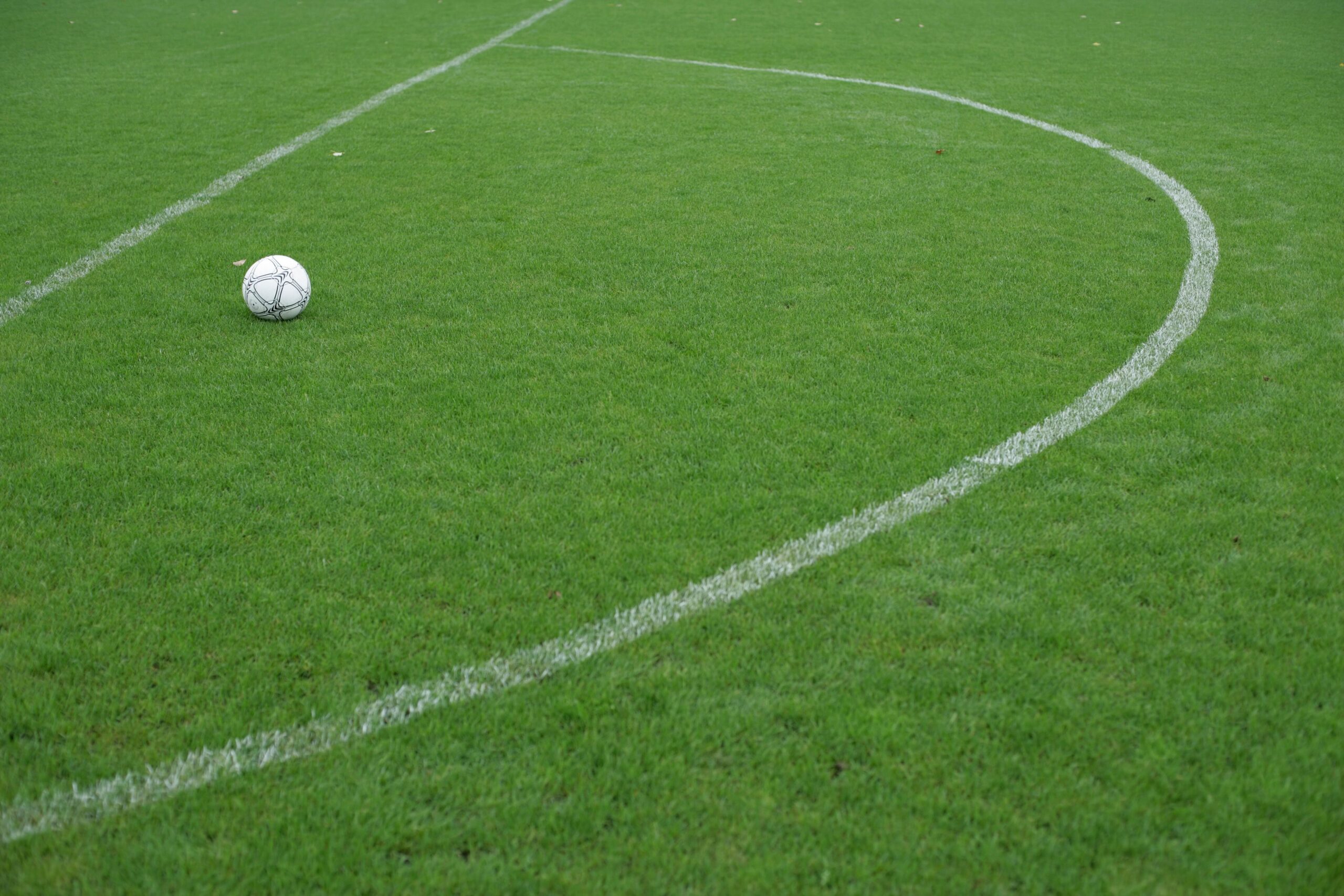 A white soccer ball sits on a mown grass pitch beside the painted white lines of the penalty area
