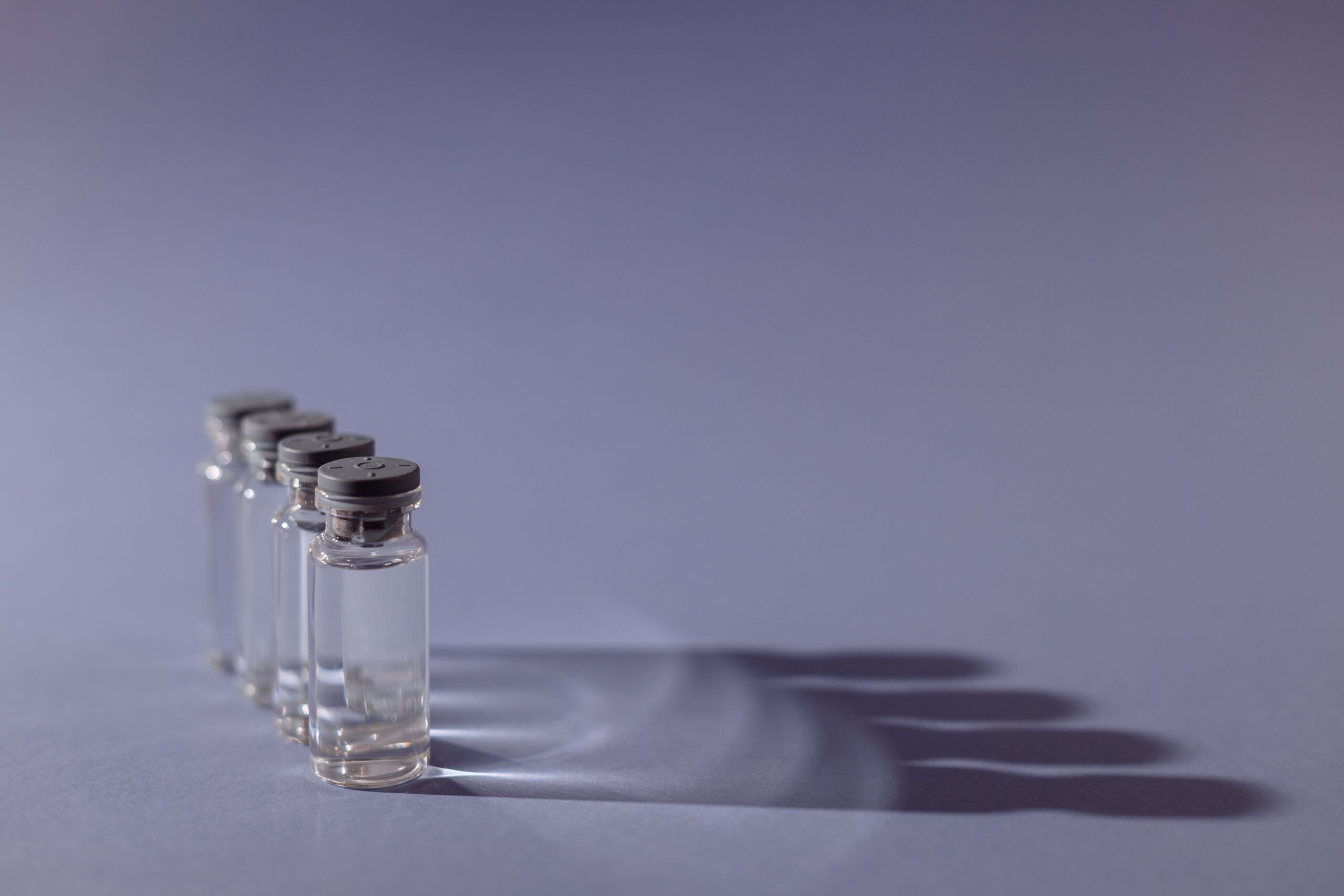 Four unlabeled glass medical vials with rubber stoppers stand in a row on a plain grey surface, casting long shadows