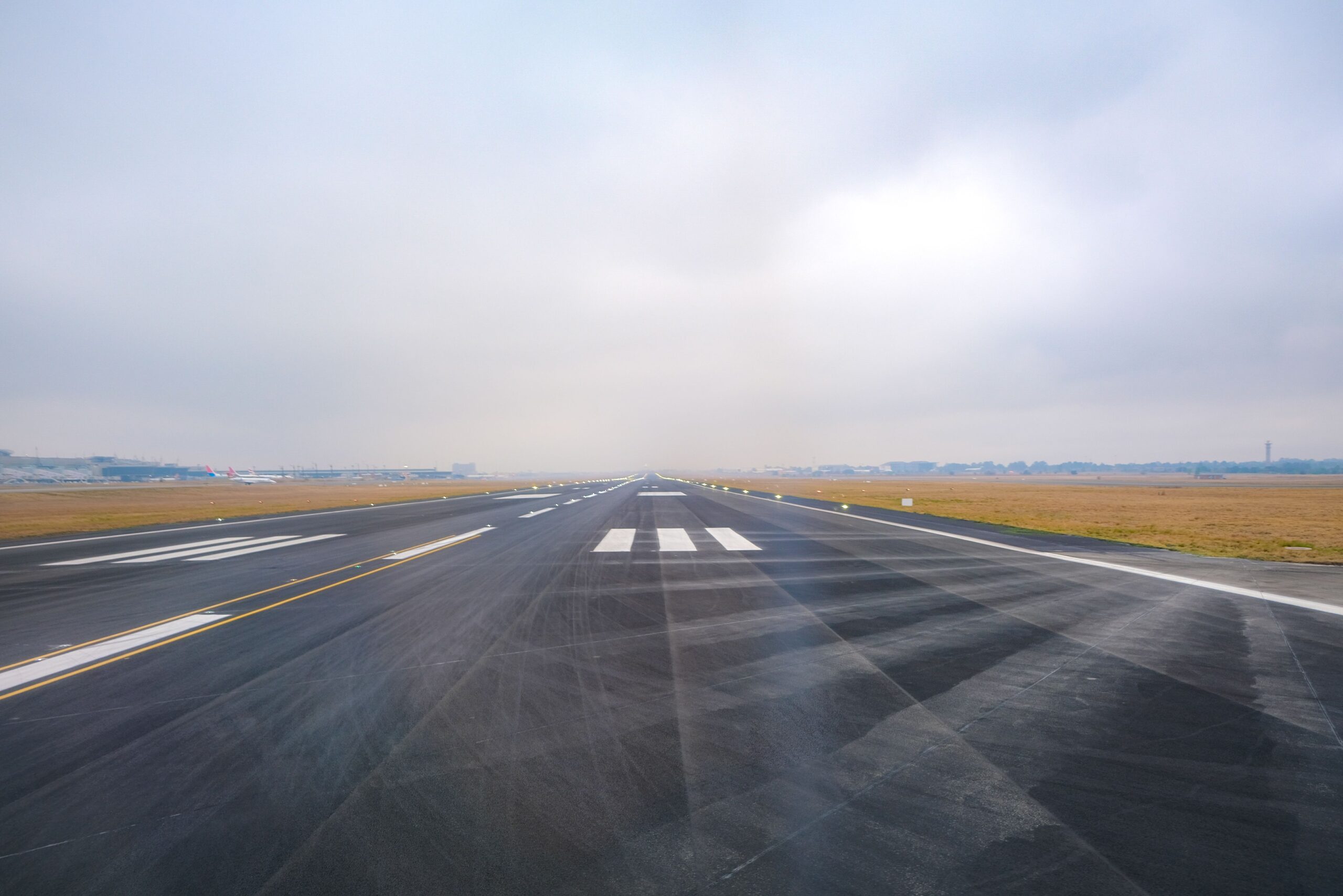 An empty airport runway seen from ground level on a hazy day, with runway markings and edge lights leading toward distant terminal buildings