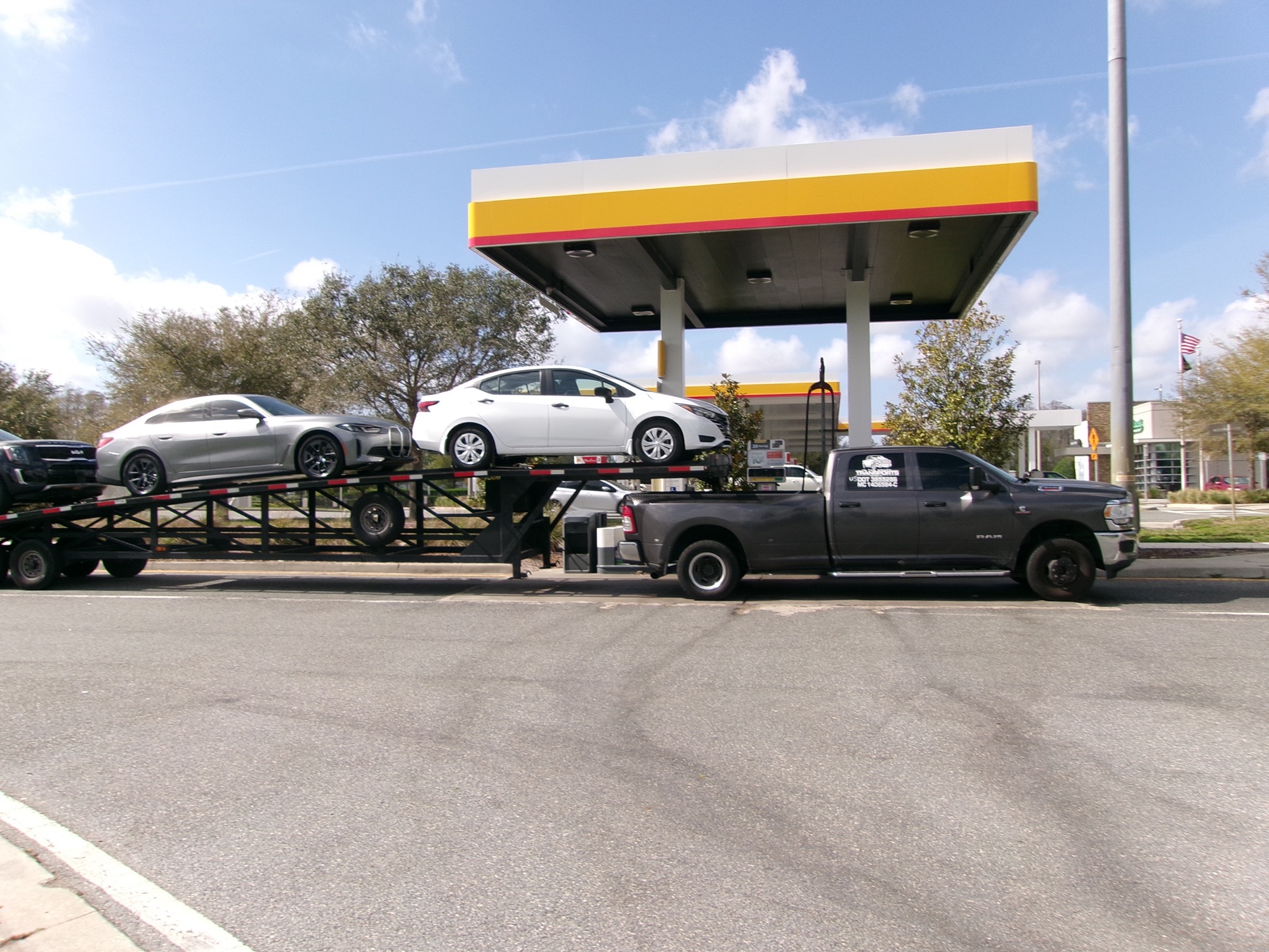 A heavy-duty pickup truck towing a trailer loaded with three cars stops under a gas station canopy at a Florida highway service plaza.
