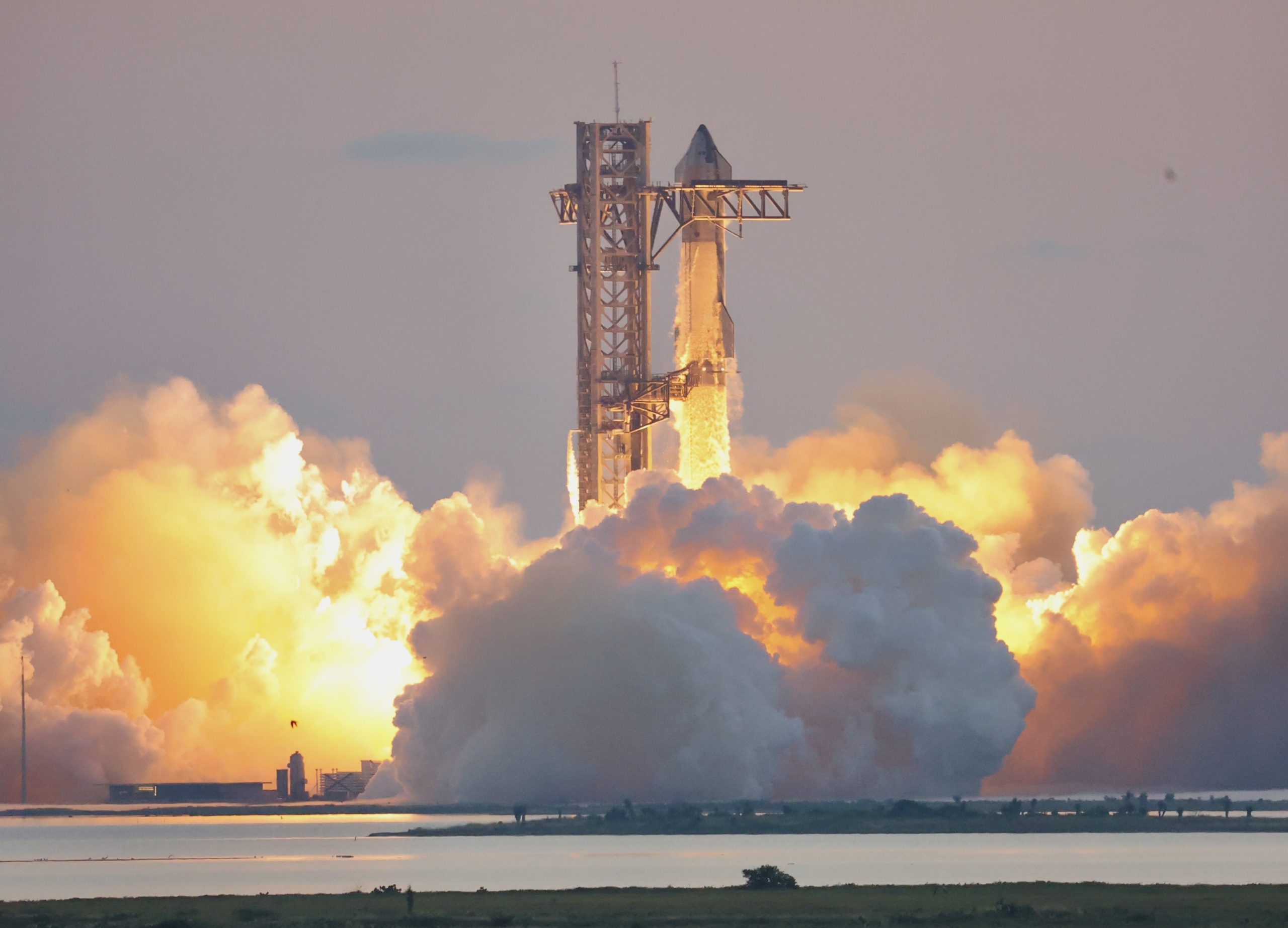 SpaceX's Starship lifting off beside its launch tower at Starbase, Texas, in a large cloud of exhaust at sunrise.
