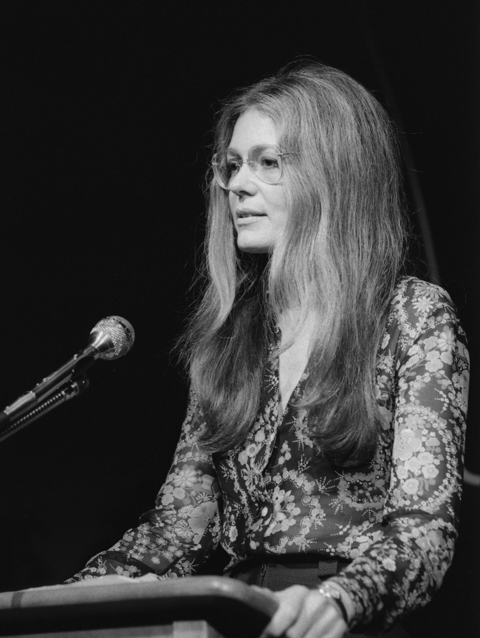 Gloria Steinem speaking at a podium microphone in 1975, in a patterned blouse, photographed in black and white