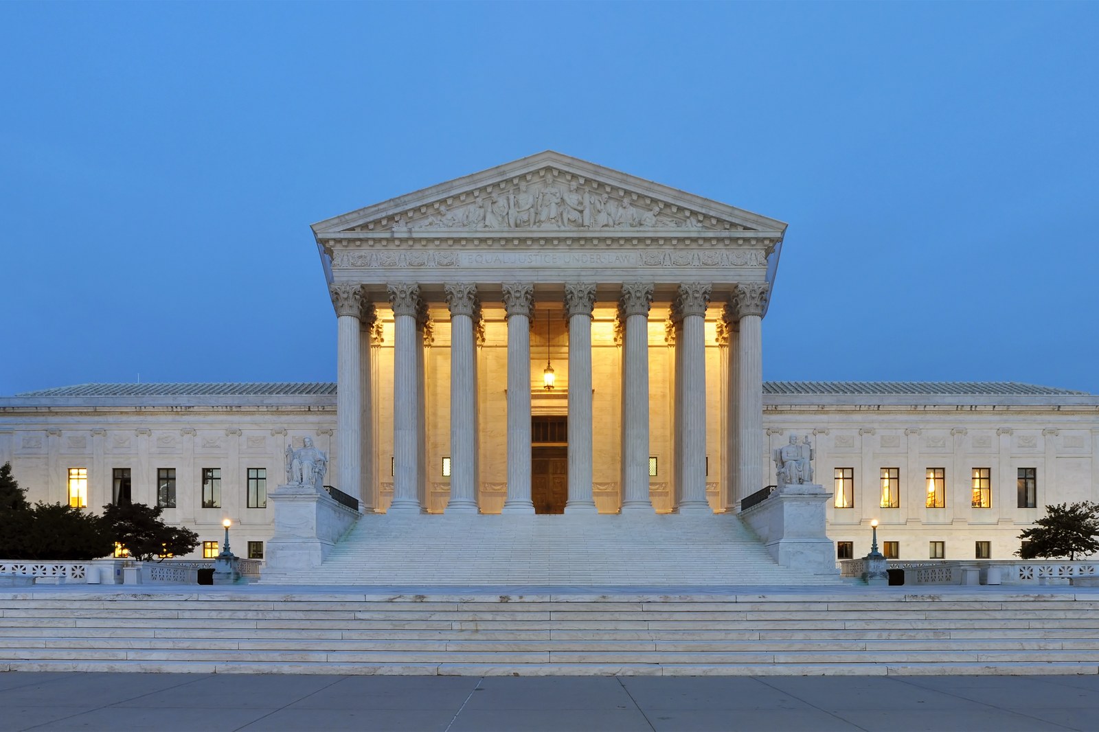 The west facade of the United States Supreme Court Building in Washington at dusk, its columns lit from within and the words 'Equal Justice Under Law' carved above them.
