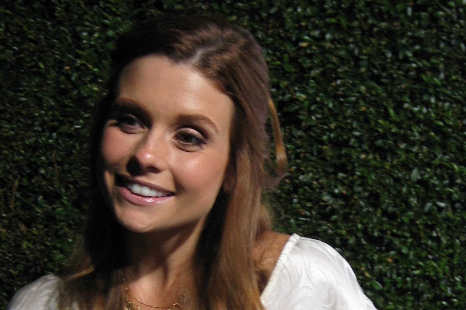 Actress JoAnna Garcia Swisher, smiling, with long auburn hair and a white top, in front of a green hedge.