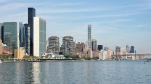 The United Nations Secretariat tower and the East River shoreline of Manhattan under a clear sky, with the Queensboro Bridge in the distance.