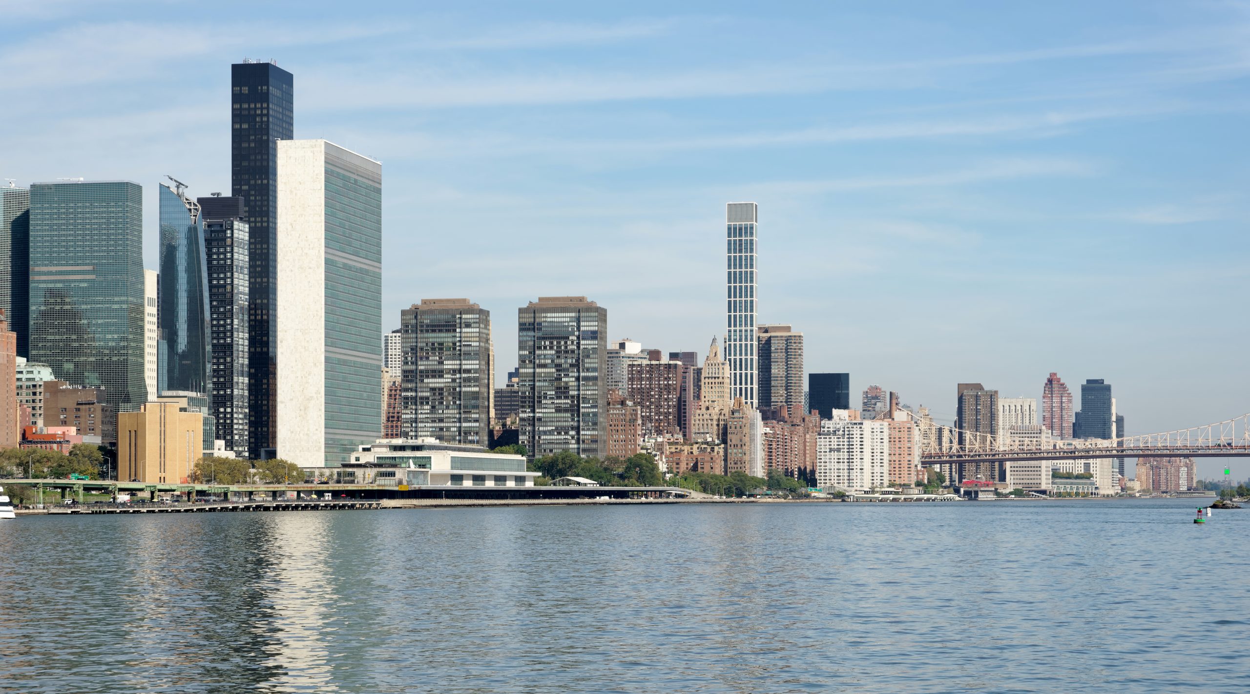 The United Nations Secretariat tower and the East River shoreline of Manhattan under a clear sky, with the Queensboro Bridge in the distance.